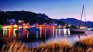 A coastal town at dusk with buildings, boats, and a sailboat on calm waters. - Vourkari Village (Kea)