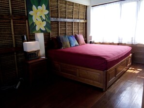 A wooden bed with a red bedspread, a nightstand with a lamp, and a wall with a large floral painting. - Bamboo bungalow (Kuta)