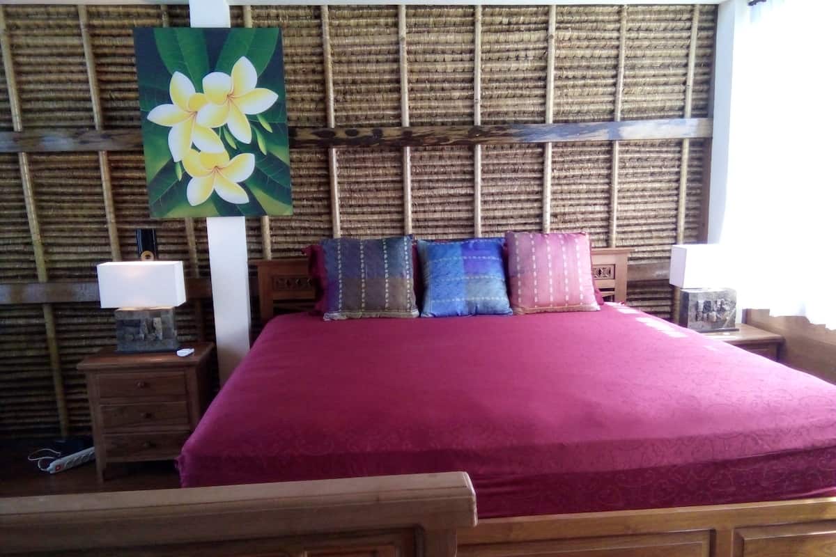 Una cama con una colcha roja, cabecero de madera y varios almohadones. Hay una mesita de noche con una lámpara y un cuadro de flores en la pared.