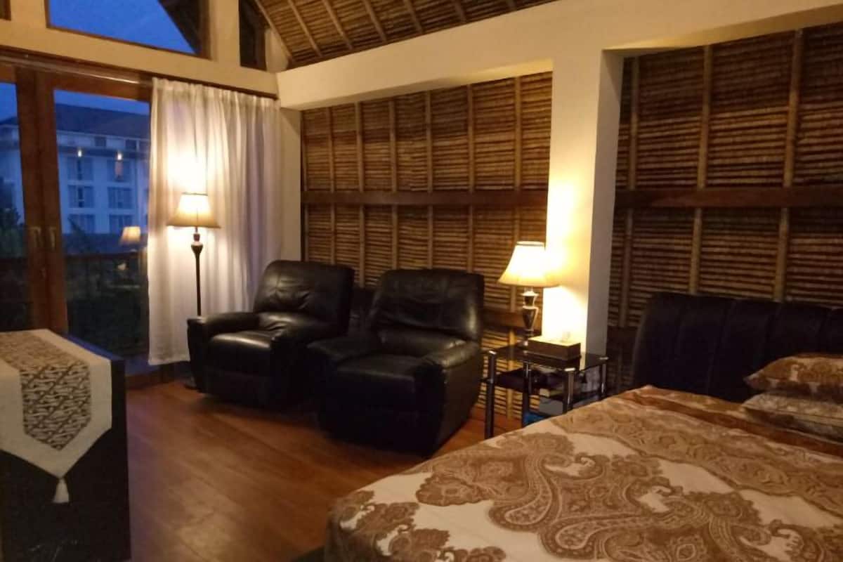 Habitación de hotel con una cama grande, dos sillones de cuero, una mesita de noche con una lámpara y vistas a edificios a través de una ventana.