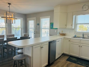 A kitchen with white cabinets, a dishwasher, a dining area with chairs, and a large window. - Long Grass 5 Bedroom Home by RedAwning (Supply)