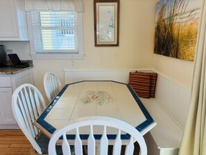 A dining area with a tiled table, white chairs, a framed picture, and a window with blinds.