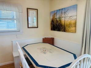 A dining area with a table, chairs, a window with blinds, a framed picture, and a large landscape painting.