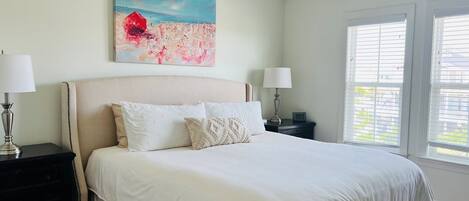 Una camera da letto con un letto, due comodini con lampade, una ventola a soffitto, una finestra con persiane e un quadro appeso al muro.