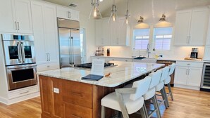 A modern kitchen with white cabinets, stainless steel appliances, a central island with chairs, and pendant lighting. - The Rainbow Connection 4 Bedroom Home by RedAwning (Supply)