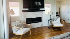 A living room with a fireplace, two white armchairs, a TV mounted above the fireplace, and a small side table with a lamp. - The Rainbow Connection 4 Bedroom Home by RedAwning (Supply)