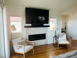 A living room with a fireplace, two white armchairs, a TV mounted above the fireplace, and a small side table with a lamp. - The Rainbow Connection 4 Bedroom Home by RedAwning (Supply)