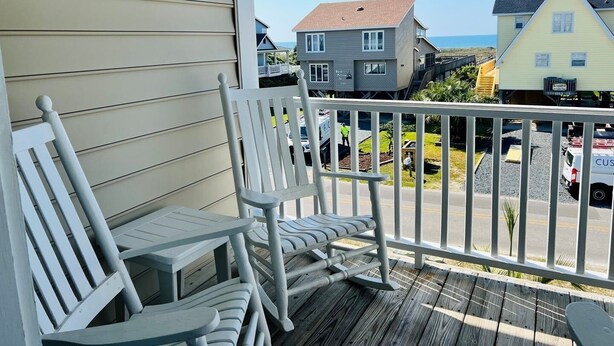 A wooden deck with white chairs, a railing, and a view of houses and a road.