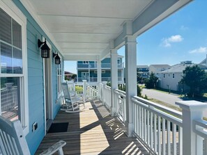 A covered porch with white railings and a wooden deck, overlooking a residential area with houses. - Blue Sunset 4 Bedroom Home by RedAwning (Supply)