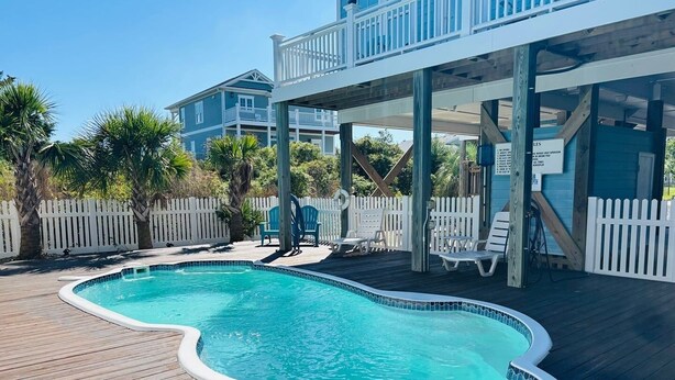 Une piscine avec une terrasse en bois, une clôture blanche et un espace abrité équipé de chaises.