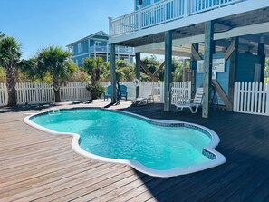 Une piscine avec une terrasse en bois, une clôture blanche et un espace abrité équipé de chaises.