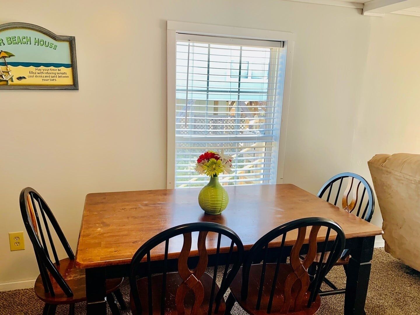 A dining area with a wooden table, four chairs, a vase with flowers, and a beach-themed sign on the wall.