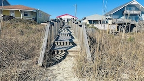 A wooden boardwalk leading to a beach house with a red roof, surrounded by dry grass and sand.