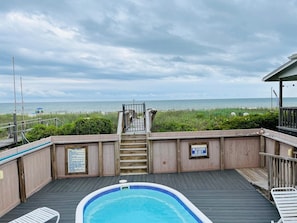 A wooden deck with a pool, a staircase leading to a beach, and a lifeguard sign.