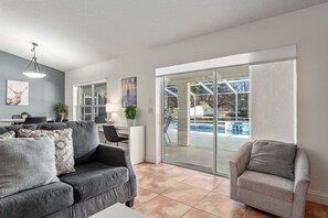 A modern living room with a grey sofa, a grey armchair, and a dining area with a table and chairs. There is a large window with a view of a swimming pool and greenery outside.