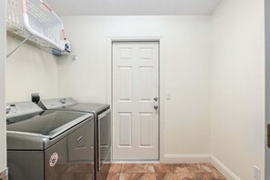 A laundry room with a front-loading washer and dryer, a white door, and a shelf above the washer. - North Timucuan Trail 1208 3 Bedroom Villa by RedAwning (Inverness)