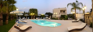 Une piscine entourée de palmiers et d’un bâtiment doté d’un balcon, avec des chaises longues et des parasols à proximité.