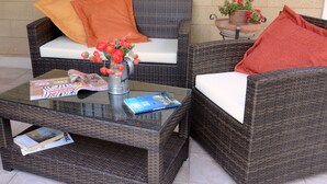 Un sofá de mimbre con cojines naranja, una mesa de centro de vidrio con revistas y un libro, y una planta en maceta con flores rojas.