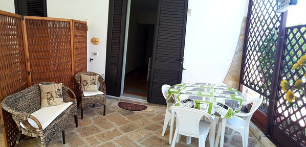 Un espace patio aménagé avec des meubles en osier, une table recouverte d’une nappe à motifs et une vue sur l’extérieur à travers une clôture à treillis.