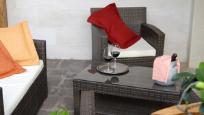 Un sofá de mimbre con un cojín blanco y almohadas naranja y rojas, una mesita con una copa de vino y una botella, y una silla de mimbre con una almohada roja.