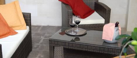 Un sofá de mimbre con un cojín blanco y almohadas naranja y rojas, una mesita con una copa de vino y una botella, y una silla de mimbre con una almohada roja.