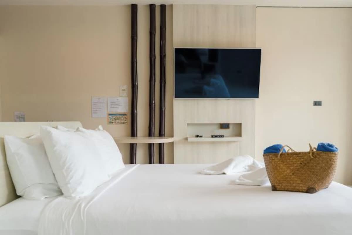 A hotel room with a bed, a television mounted on the wall, a small shelf, and a wicker basket with towels.