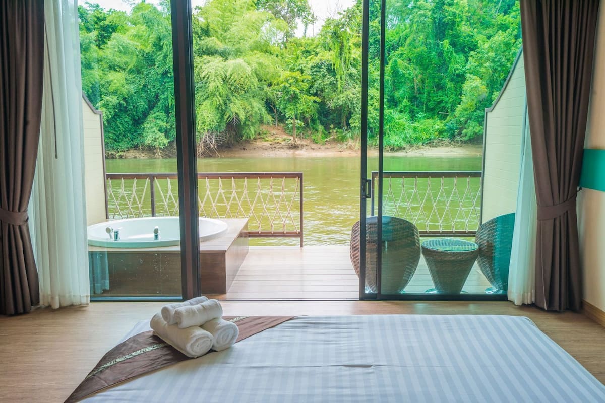 A room with a bed, a bathtub, and a view of a river and forest.
