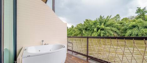 Un balcon avec une baignoire blanche, une terrasse en bois et une vue sur une rivière bordée d’une végétation luxuriante.