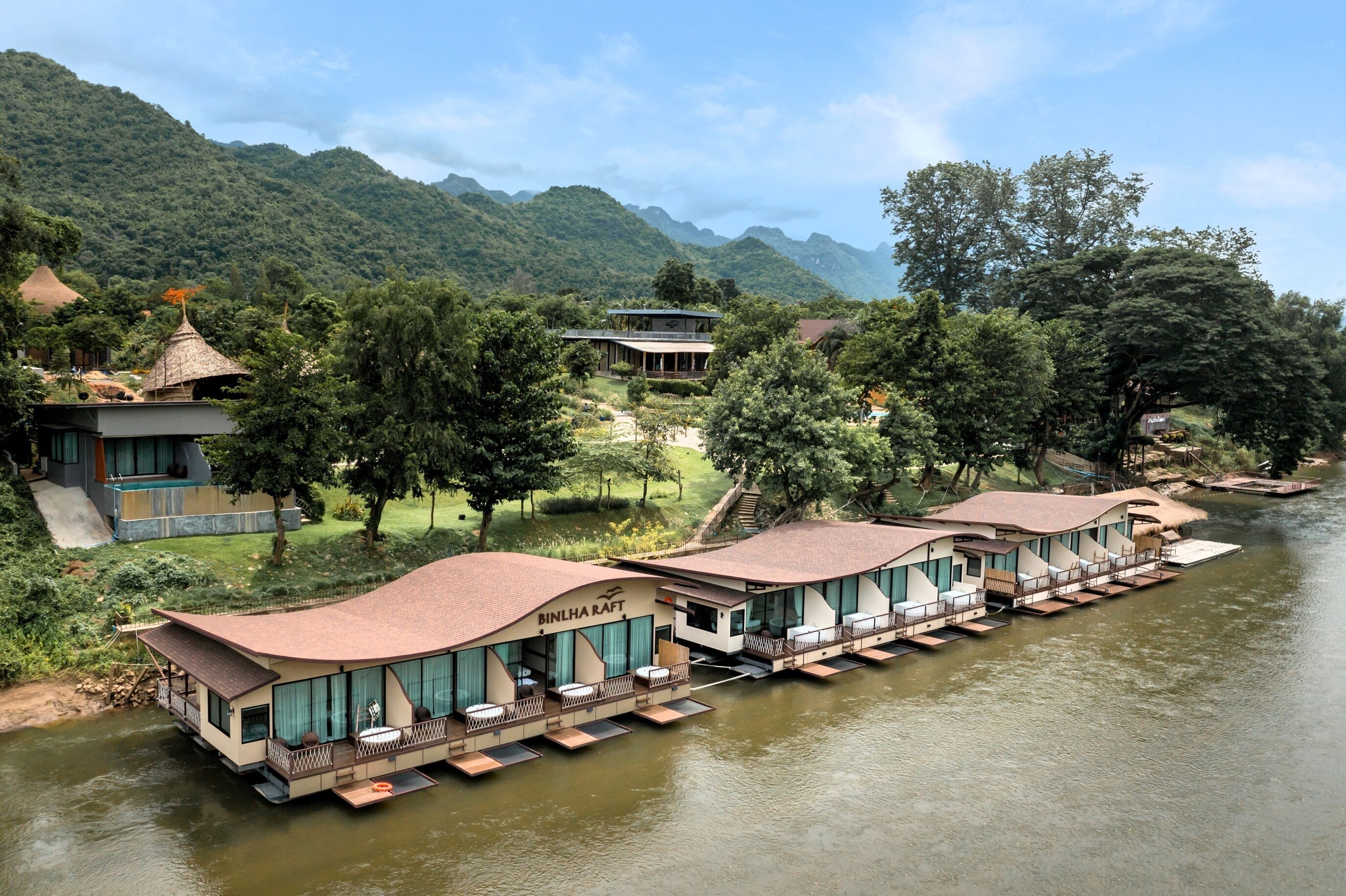 Un hotel junto al río con alojamientos flotantes, una piscina y montañas al fondo.