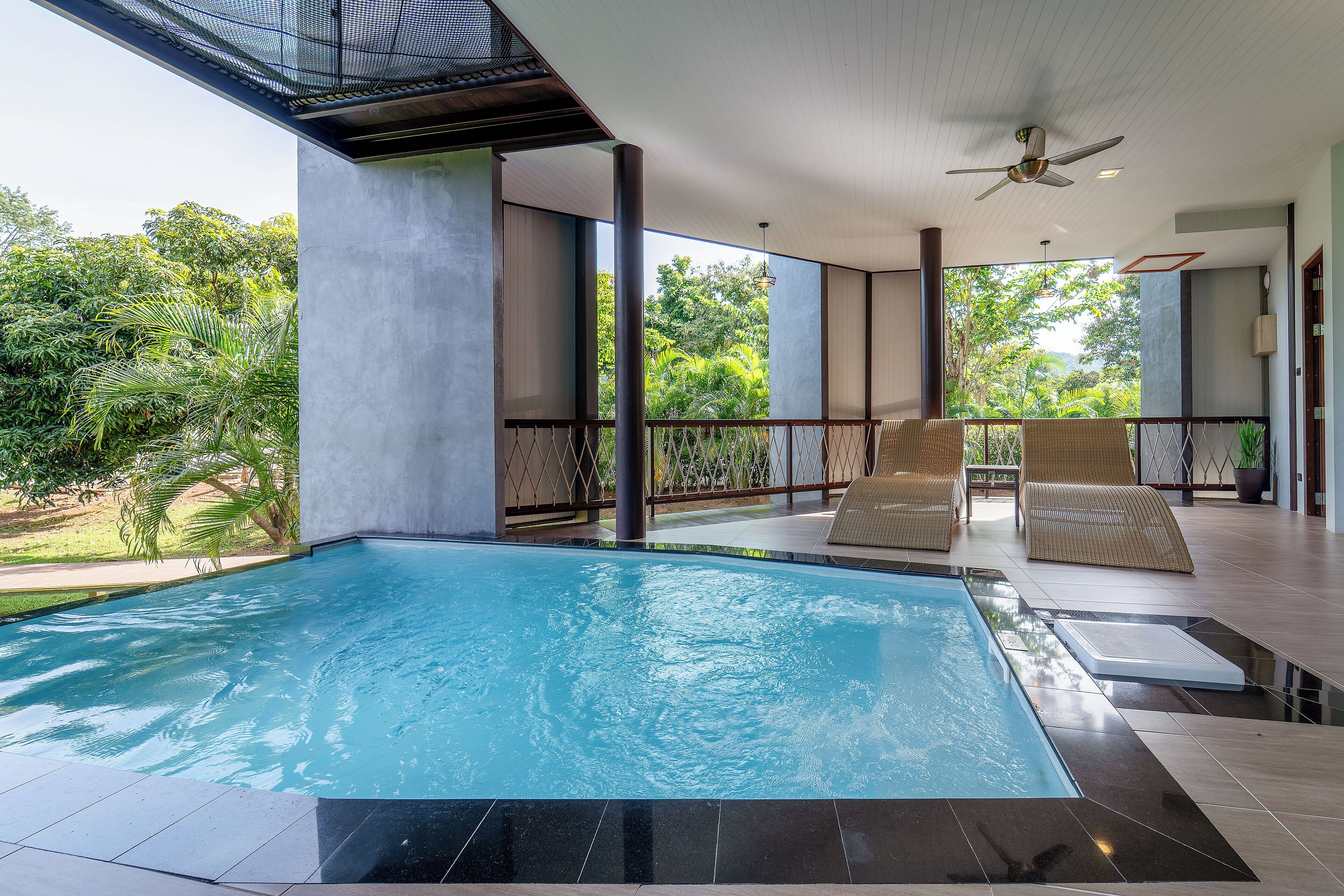 A modern indoor swimming pool with clear glass walls, surrounded by lounge chairs and a ceiling fan.