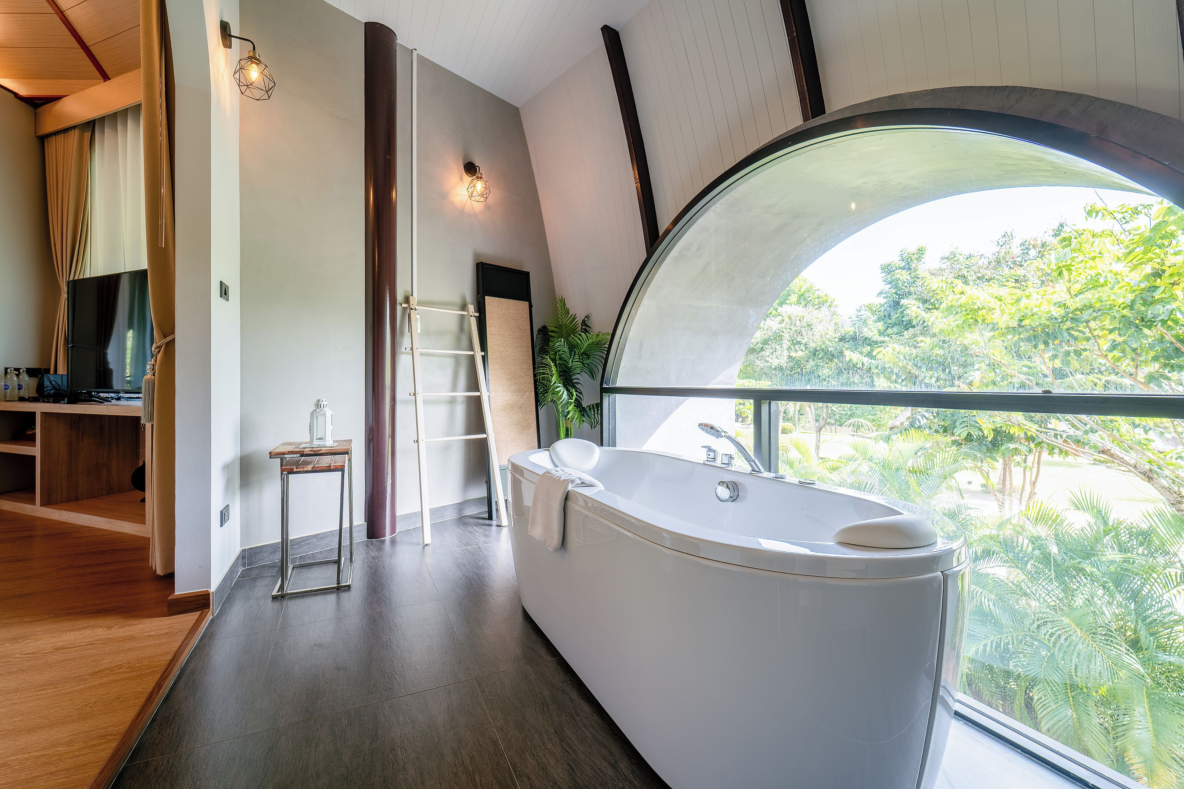 A modern bathroom with a large bathtub, a wooden floor, a TV, and a view of greenery through a large window.