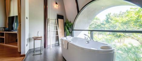 A modern bathroom with a large bathtub, a wooden floor, a TV, and a view of greenery through a large window.