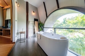 A modern bathroom with a large bathtub, a wooden floor, a TV, and a view of greenery through a large window. - Binlha Raft Resort (Sai Yok)