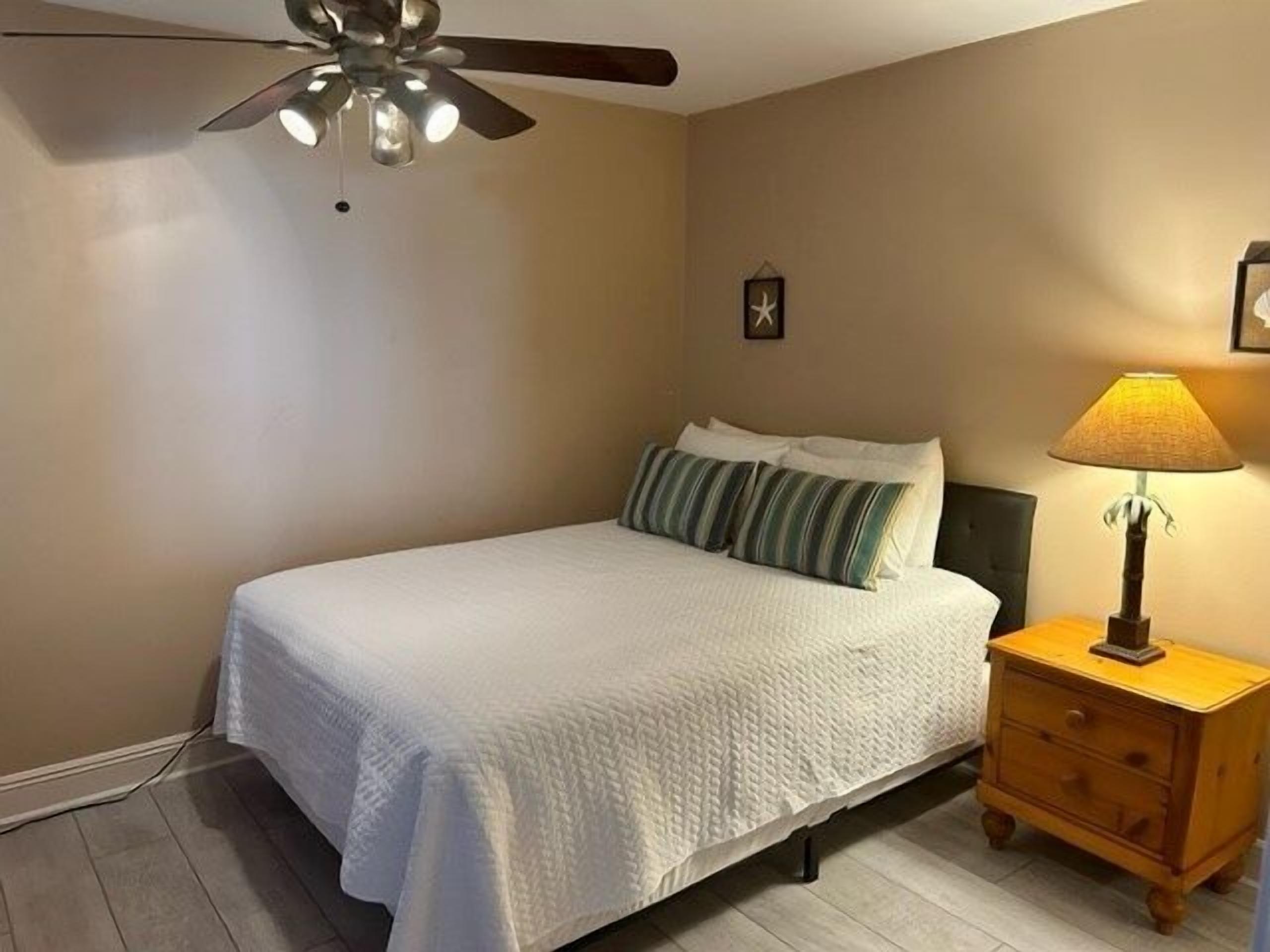 Un dormitorio con cama, mesita de noche, lámpara, ventilador de techo y un reloj de pared.