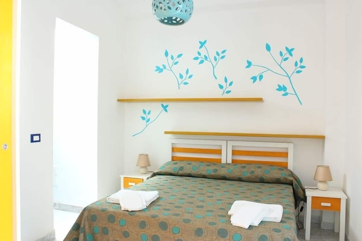 Habitación de hotel con una cama, dos mesitas de noche, una lámpara en cada una y un adorno de ramas en la pared.