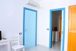 Una habitación con puerta azul, una silla blanca, un escritorio con computadora y una cocina visible a través de una puerta abierta.
