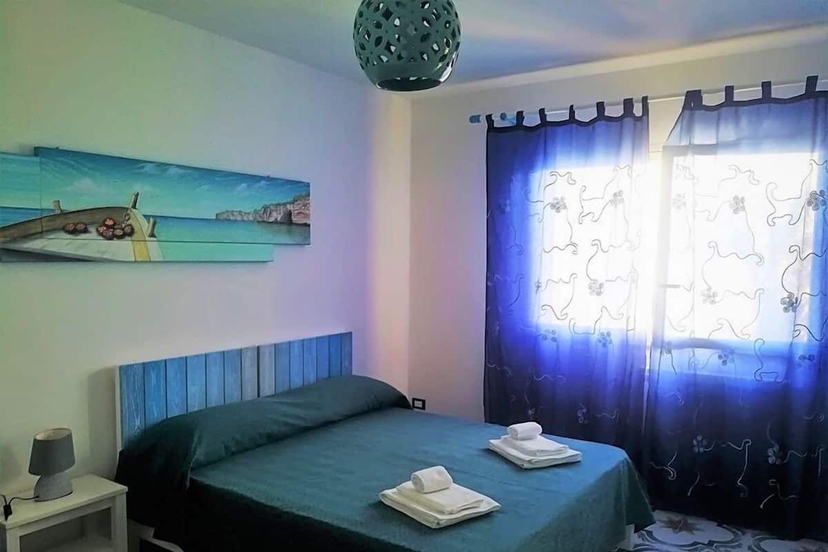 Un dormitorio con cama, mesita de noche, lámpara, un cuadro de una escena costera y una ventana con cortinas.