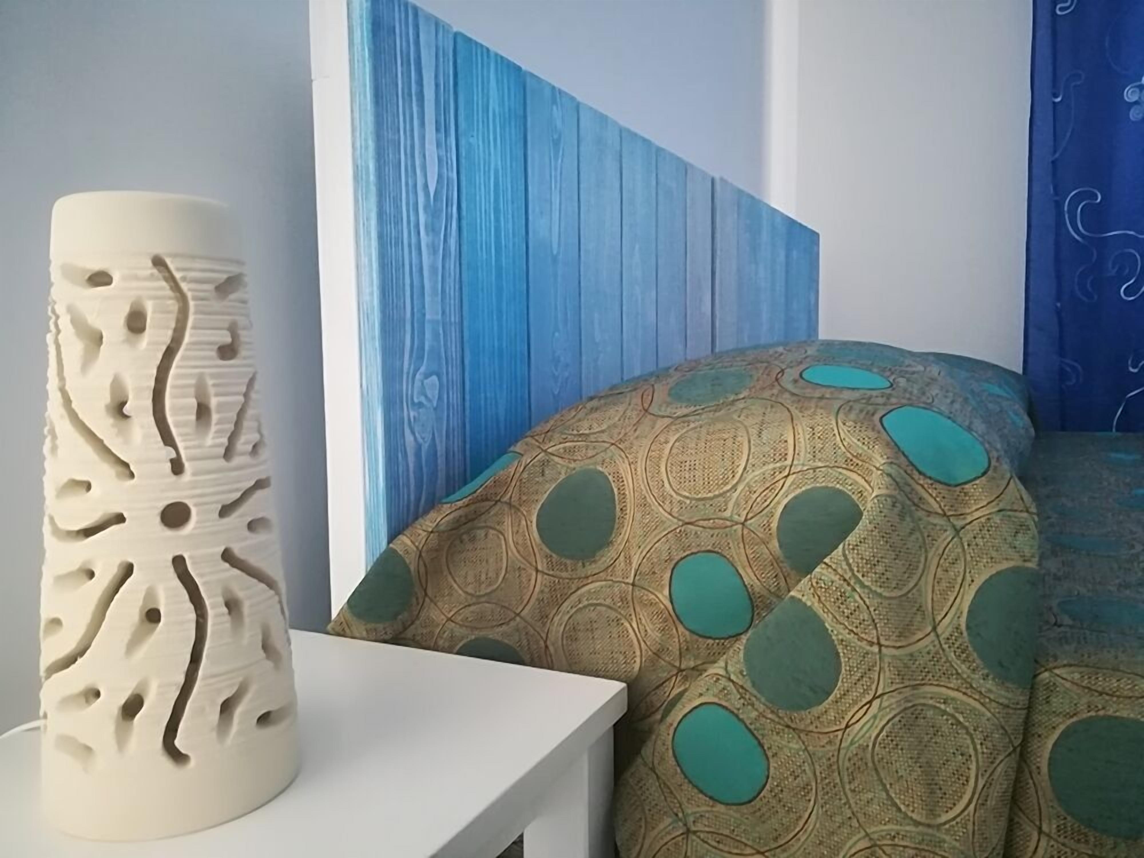 Una habitación con una mesita de noche blanca, un jarrón decorativo, una colcha estampada y una pared azul con líneas verticales.
