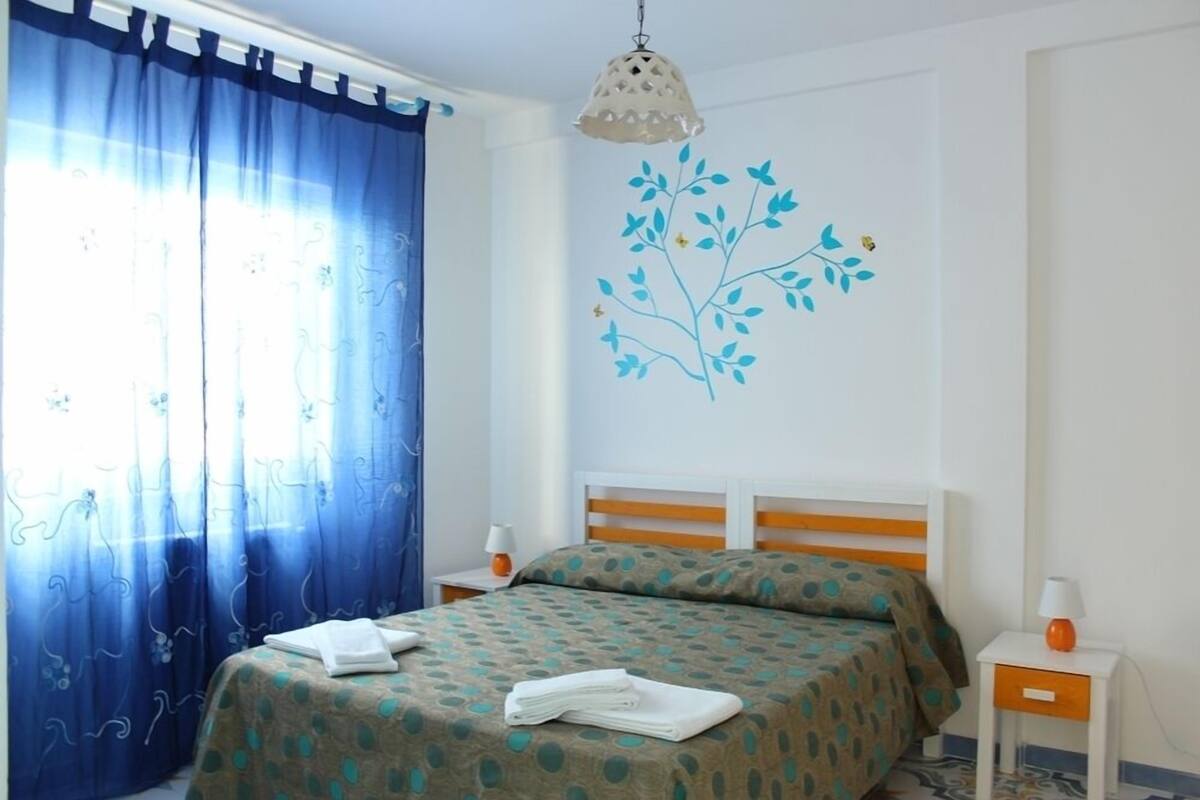 Un dormitorio con cama, cortinas, un adorno en la pared y mesitas de noche.