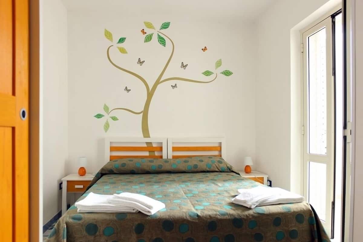 Un dormitorio con cama, mesita de noche, ventana y un adorno de pared con un árbol y mariposas.