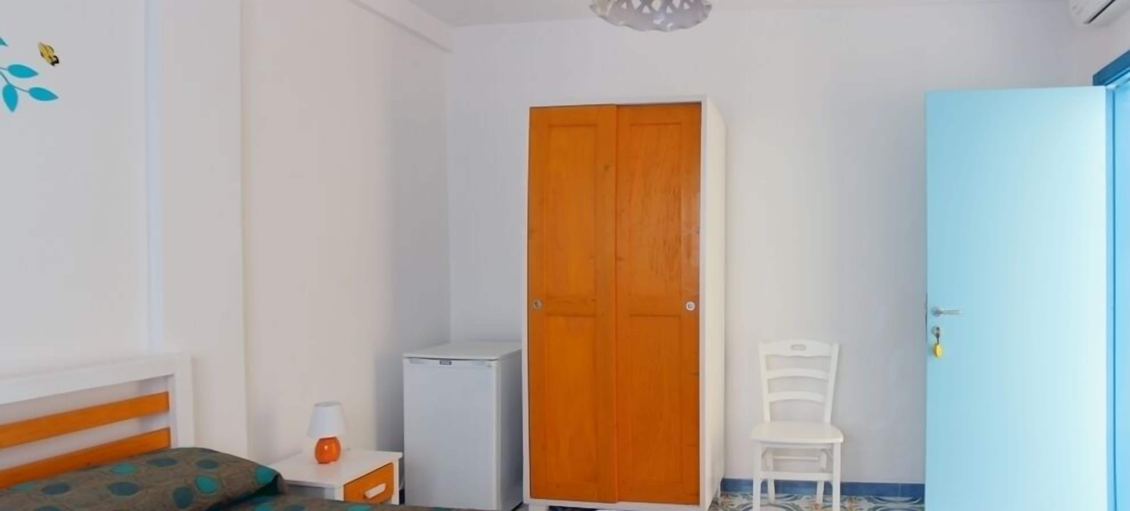 Een slaapkamer met een bed, een kledingkast, een klein tafeltje met een lamp en een stoel.