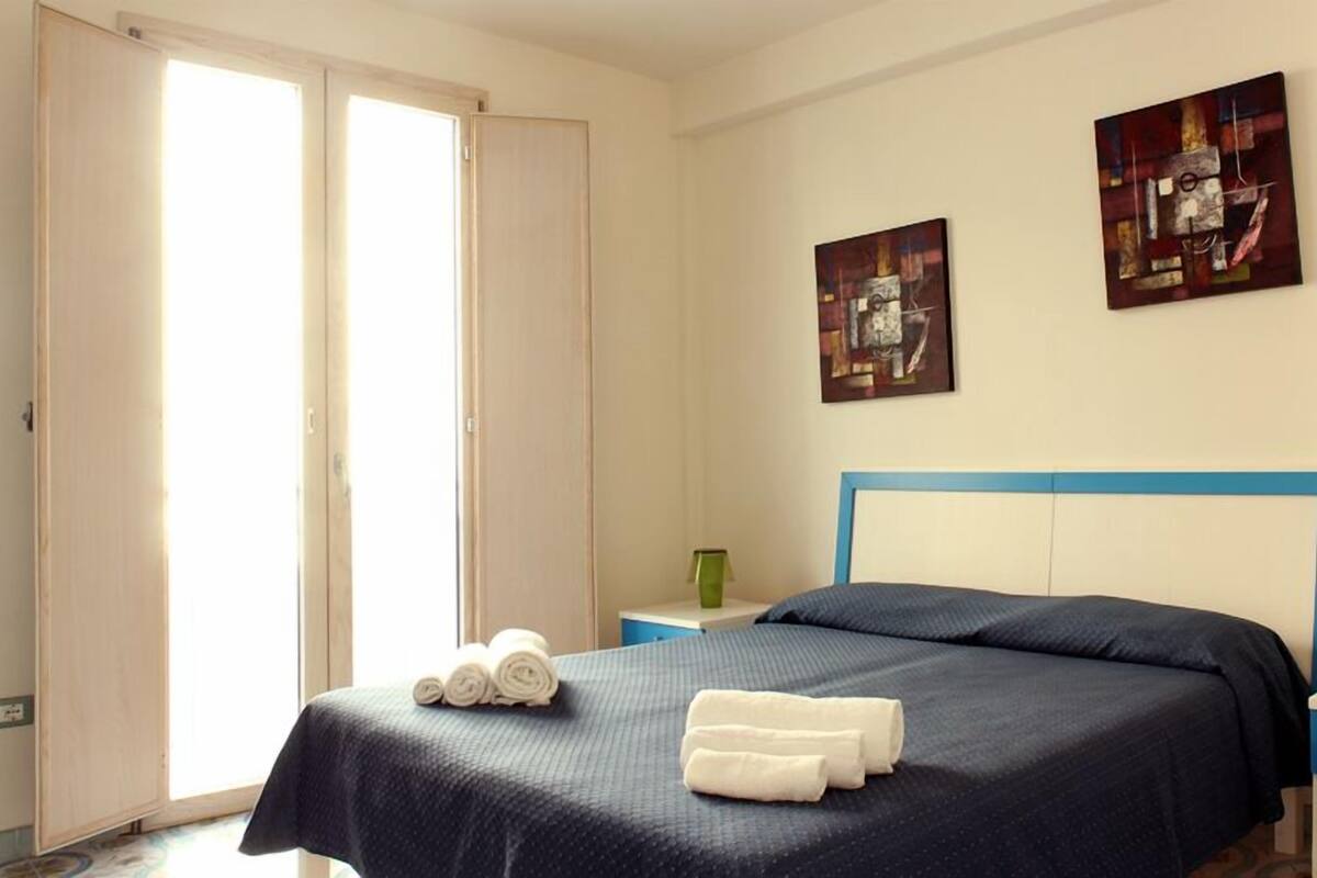 A hotel room with a bed, two paintings on the wall, a nightstand with a lamp, and a door leading to another room.