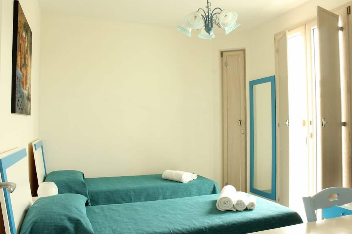 A hotel room with two beds, a painting on the wall, a chandelier, and a door with a blue frame.