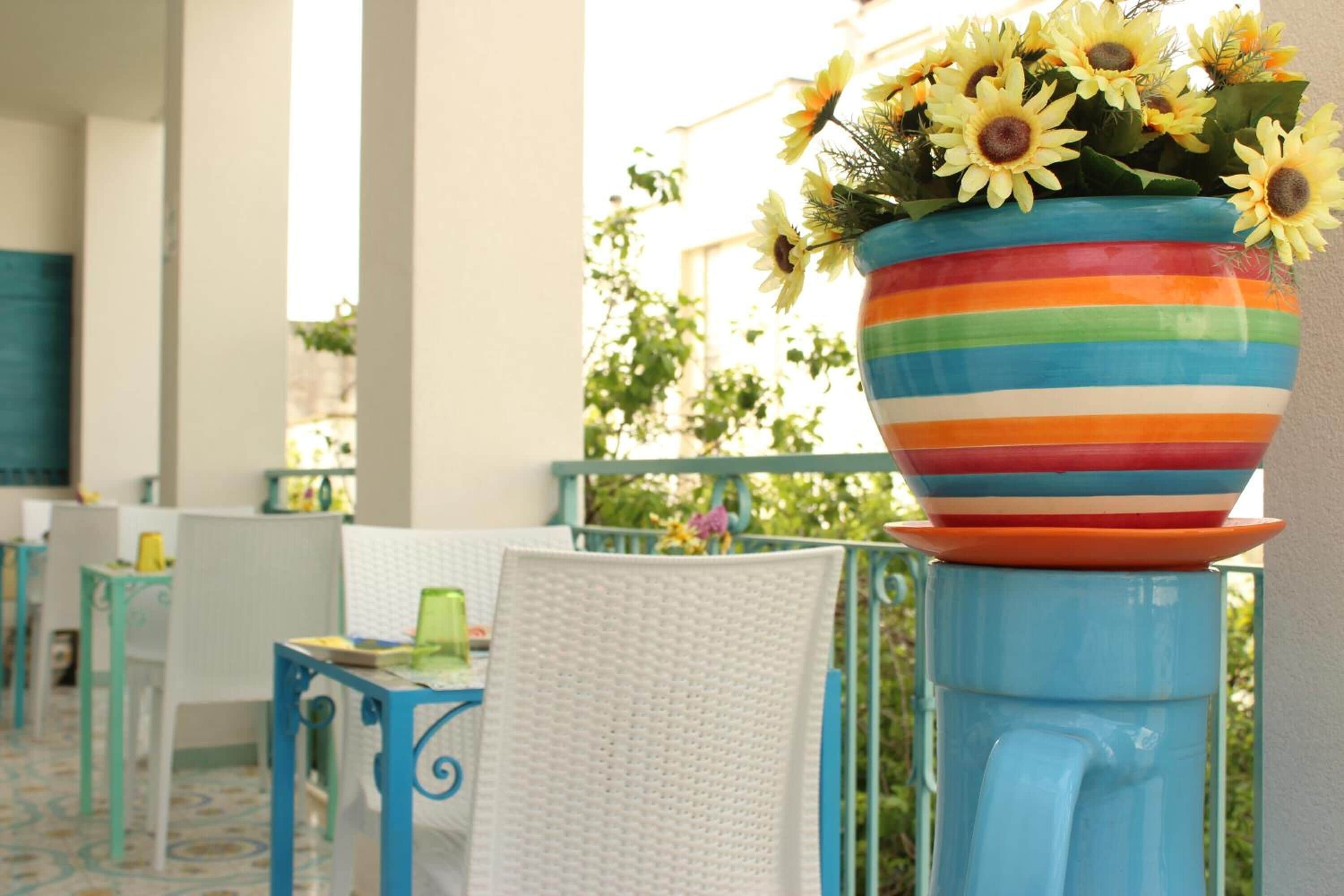 A colorful striped flower pot on a blue pedestal, placed on a balcony with white wicker chairs and a table.