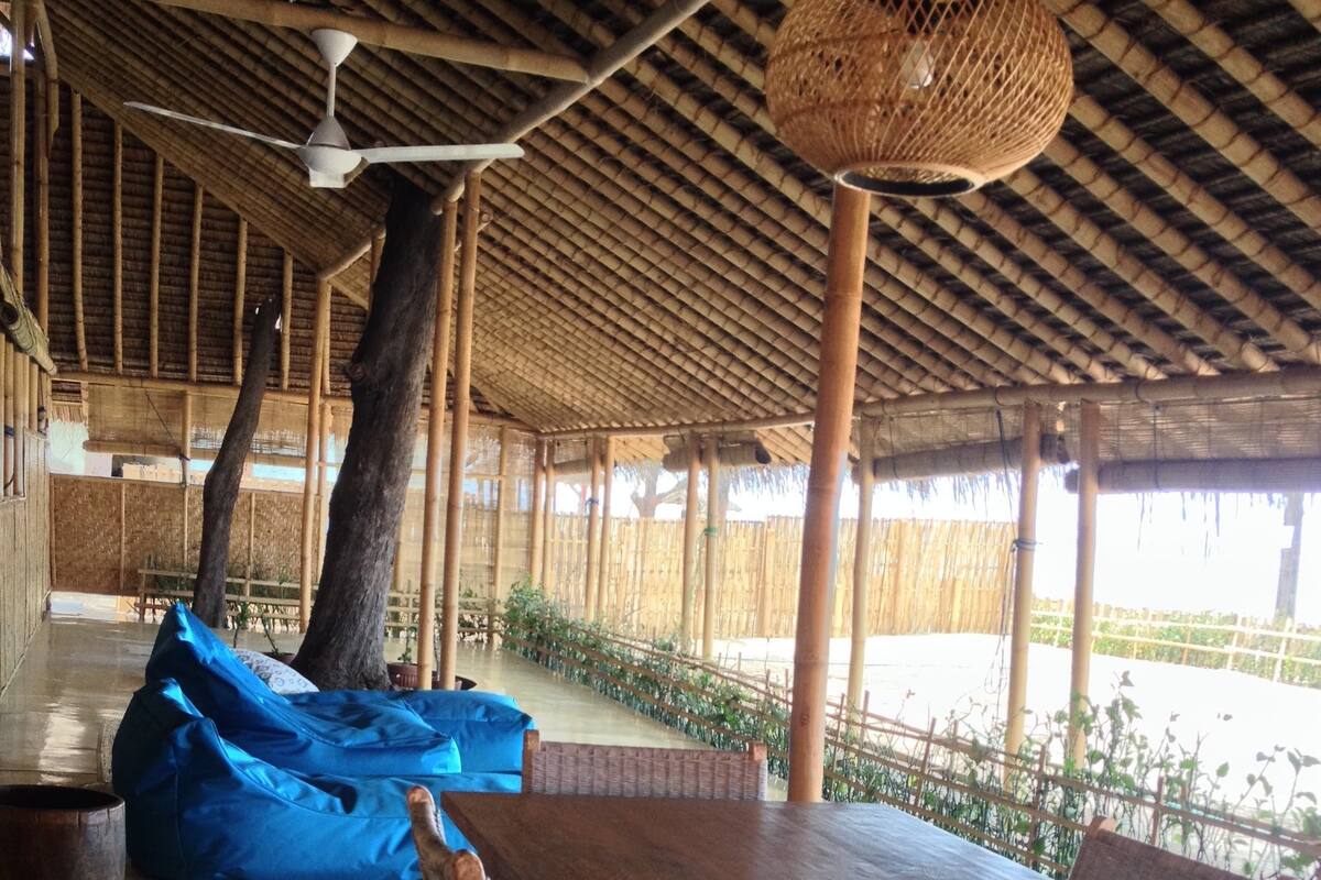 A covered outdoor dining area with a wooden table and chairs, a blue bean bag, and a hanging light fixture.