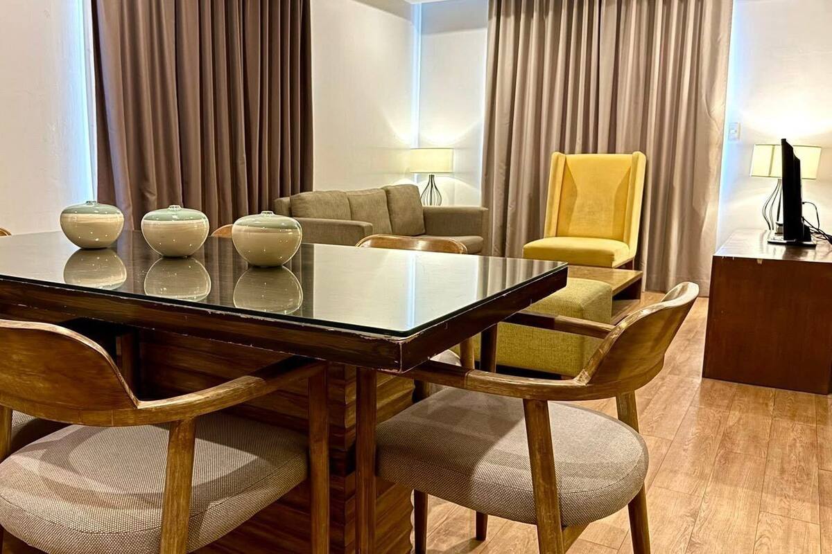 A hotel dining area with a glass-top table surrounded by wooden chairs, a yellow armchair, and a sofa.