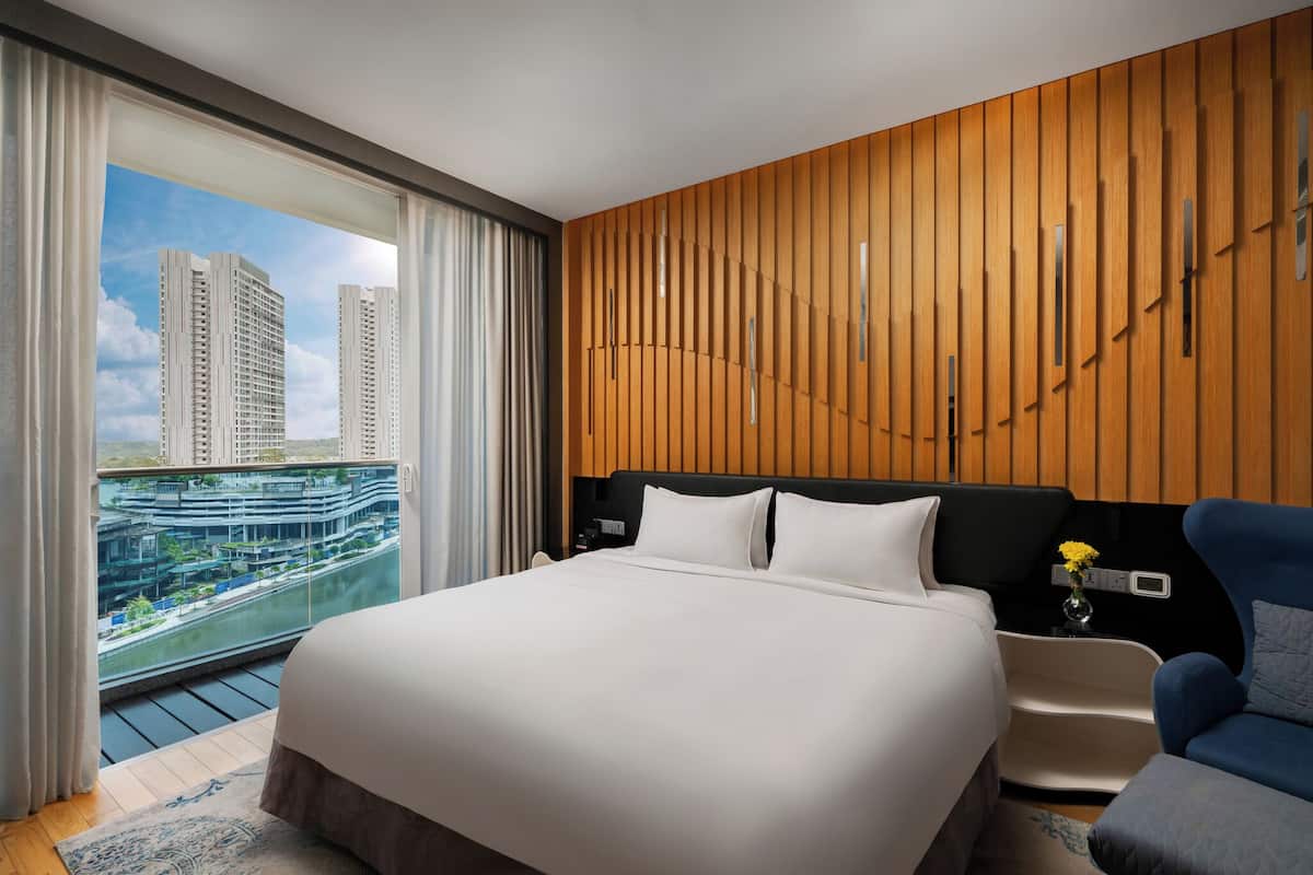 A hotel room with a large bed, a nightstand, a chair, and a view of tall buildings through large windows.