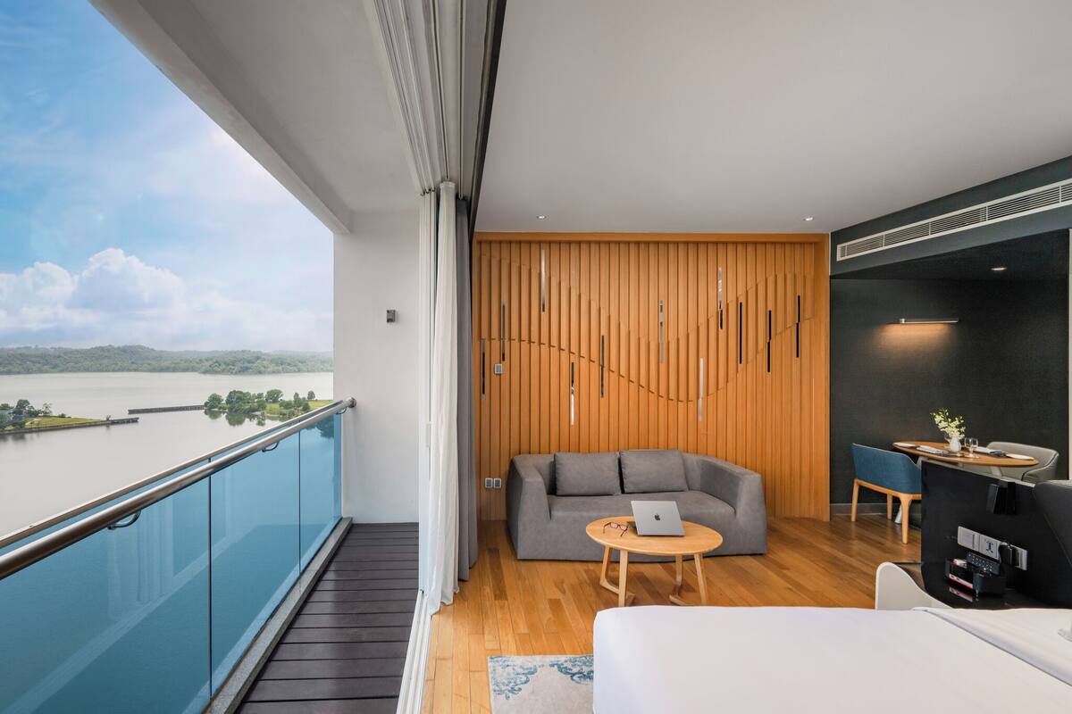 A modern hotel room with a balcony, a grey sofa, a small round table with a laptop, and a bed.