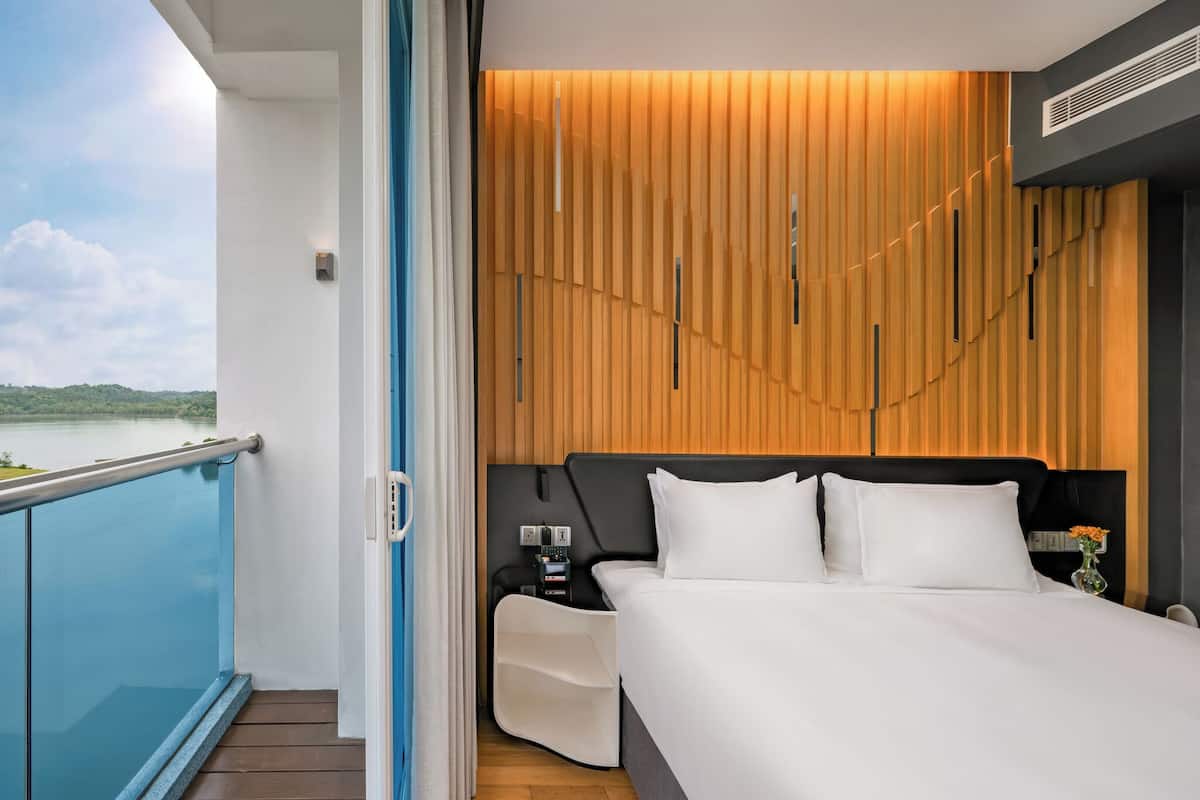 A modern hotel room with a large bed, a wooden panel wall, a nightstand with a lamp, and a view of a body of water.