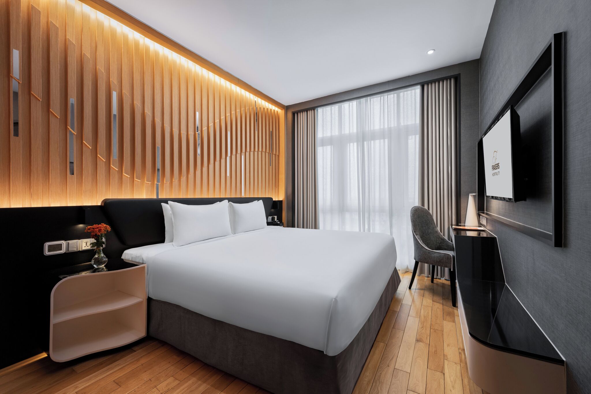 Una habitación de hotel moderna con una cama grande, un televisor de pantalla plana en la pared, una mesita de noche con un jarrón de flores y un diseño de pared de paneles de madera.